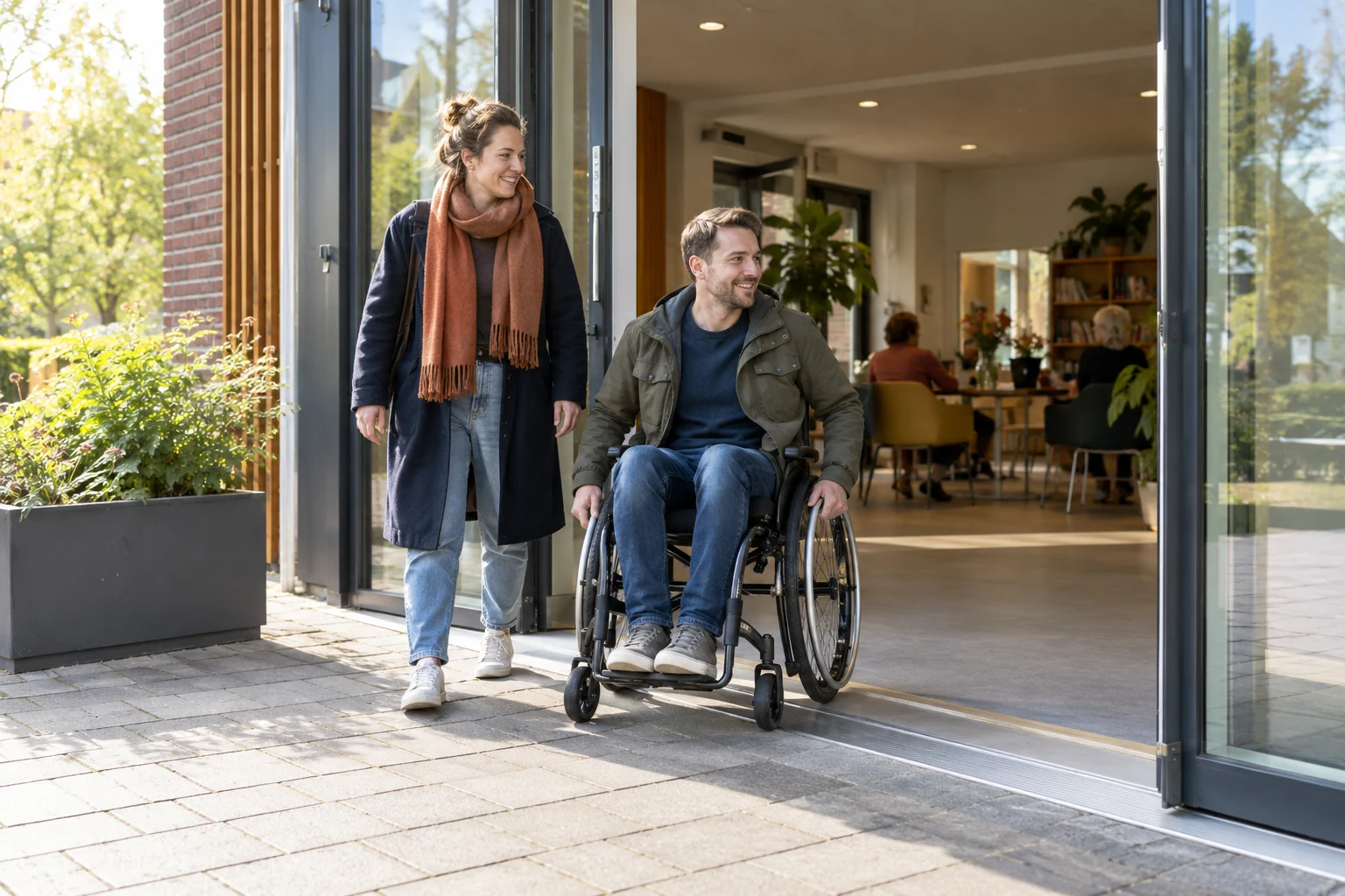 Een rolstoelgebruiker en een buurtgenoot bij de brede, drempelarme ingang van een ontmoetingsplek.