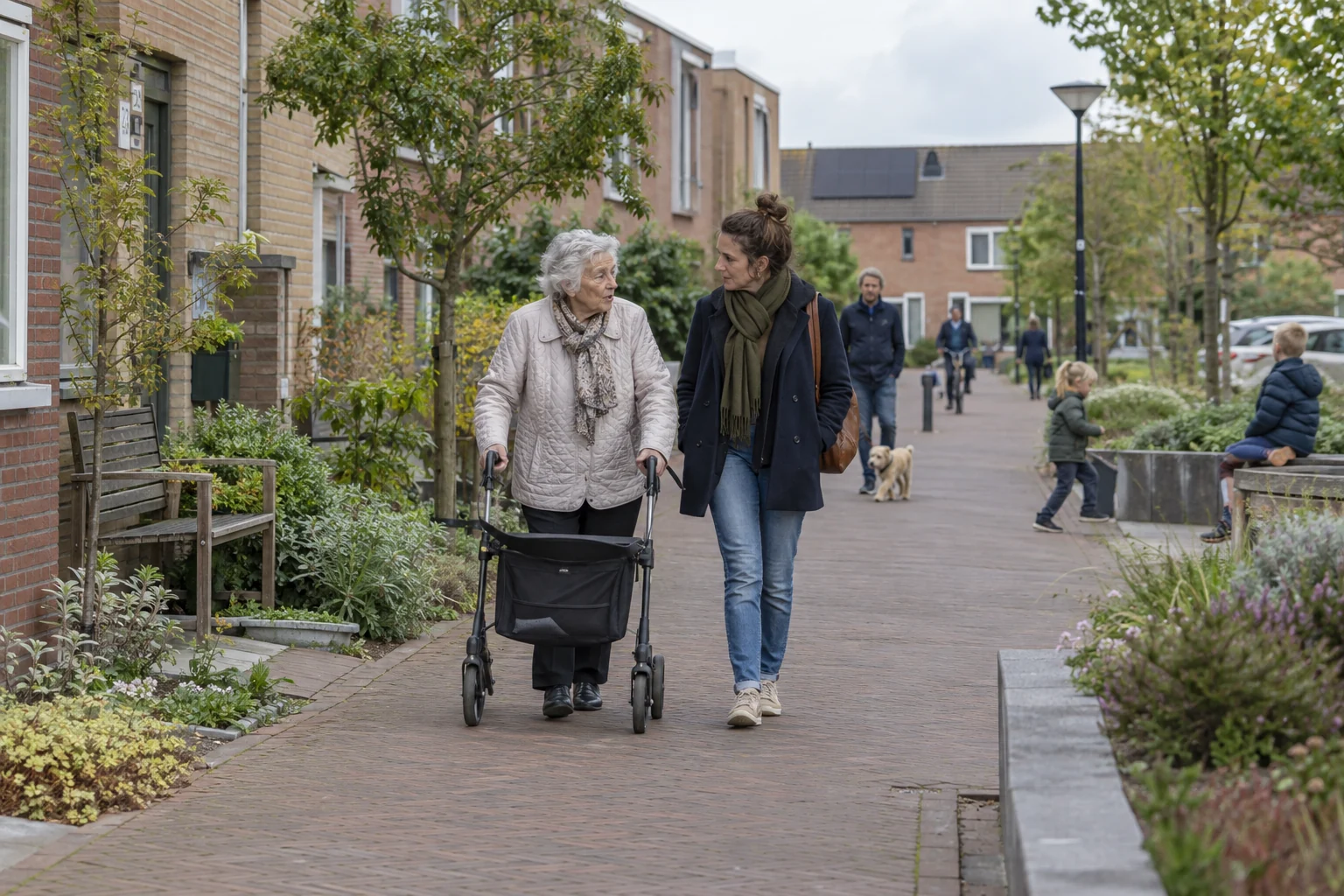 Een oudere vrouw met rollator en haar gezelschap lopen door een groene woonstraat met ruimte voor ontmoeting.