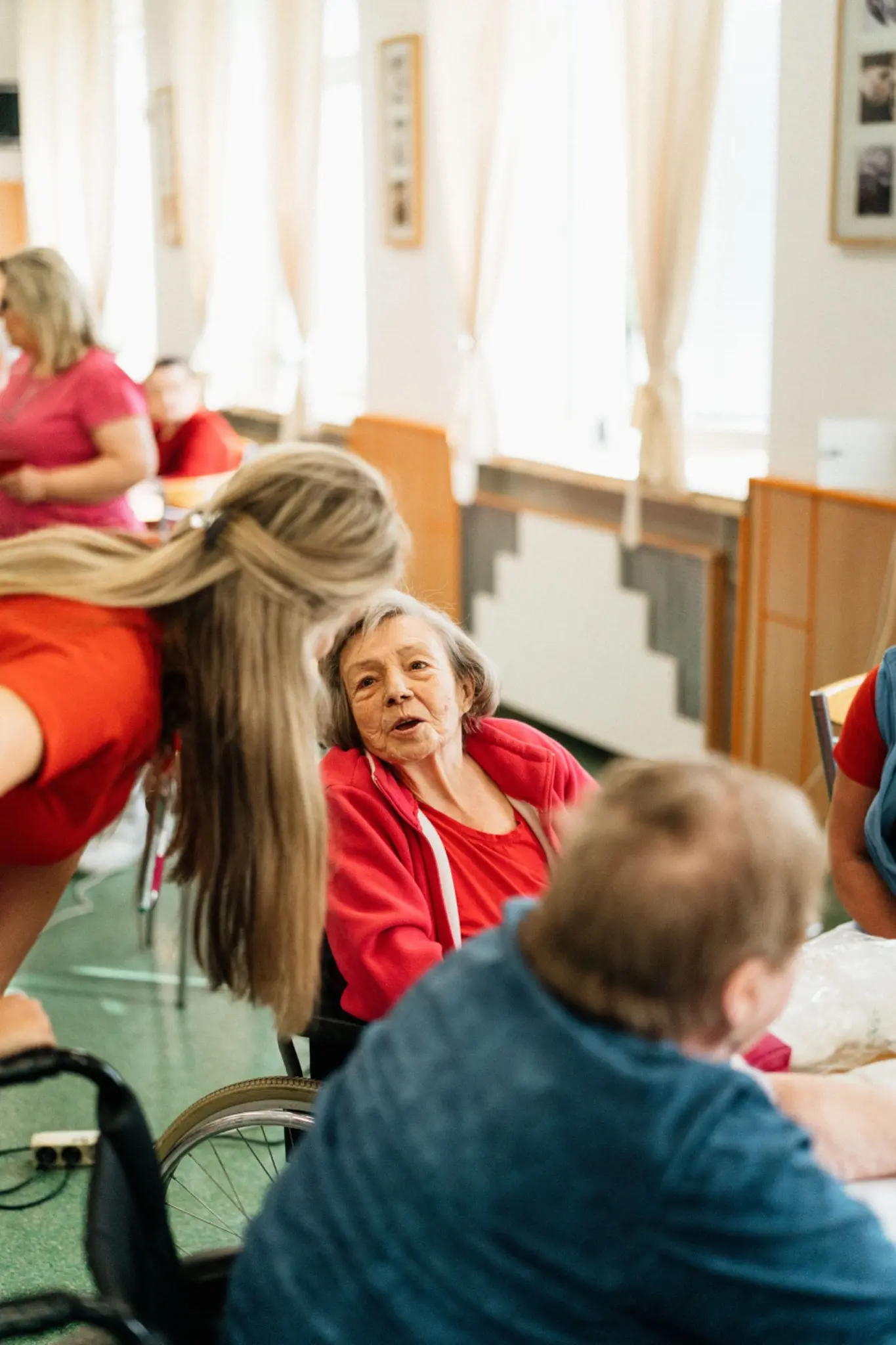 Een oudere vrouw in een rolstoel is in gesprek met andere aanwezigen tijdens een gezamenlijke activiteit.
